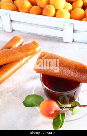 Aprikosenpastille und Aprikosenzweig mit einer Tasse Tee auf weißem Hintergrund.EINE große Anzahl von Aprikosen in einer weißen Holzkiste im Hintergrund. Natürliches und diätetisches Produkt. Kalorienarme Mahlzeiten und Snacks. Vertikales Foto. Stockfoto