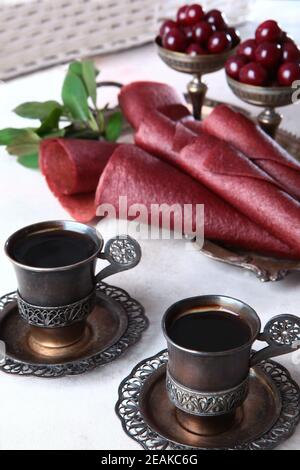Kirschpastilien werden zu einem Kegel gerollt. Zwei Tassen Kaffee in Vintage-Geschirr. Vertikales Foto. Kirschen und Äpfel sind in einer Schüssel mit Nickel-Silber. Stockfoto