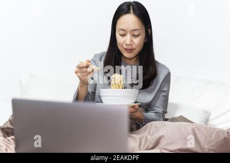 Geschäftsfrau essen Instant Nudel im Bett während der Arbeit auf Laptop zu Hause. Stockfoto