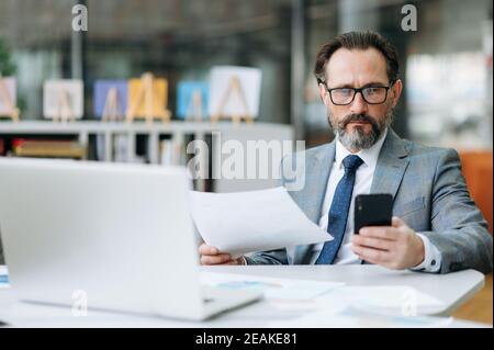 Serious mittleren Alters männliche Mitarbeiter sitzt am Tisch in modernen Büro, arbeitet mit Dokumenten. Konzentrierter Geschäftsmann ist mit Smartphone, Surfen im Internet, auf der Suche nach Ideen für Projekt oder Startup Stockfoto