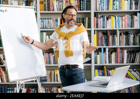 Ein bärtiger männlicher Mitarbeiter oder Lehrer mittleren Alters, der in der Nähe des Flipchart im modernen Büro steht, führt eine Online-Lektion oder Präsentation durch. Zufriedener Geschäftsmann stilvoll auf Video-Meeting oder Lektion gekleidet Stockfoto