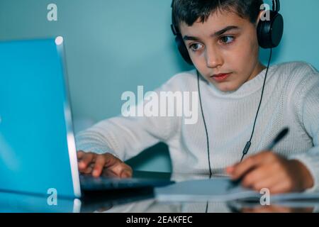Das Kind nutzt Laptop-Computer zu Hause. Er trägt Kopfhörer und verwendet ein Notizbuch. Horizontales Foto Stockfoto