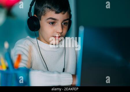 Das Kind nutzt Laptop-Computer zu Hause. Er trägt Kopfhörer und verwendet ein Notizbuch. Horizontales Foto Stockfoto
