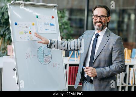 Erfolgreiche Geschäft männlich macht Präsentation auf Online-Konferenz. Ein selbstbewusster reifer Mann im formellen Anzug schaut lächelnd auf die Kamera und steht in der Nähe des Flipchart im modernen Büro Stockfoto