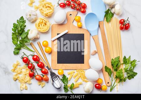 Kleine Tafel mit Kopierplatz umgeben von Zutaten zum Kochen verschiedene Arten von Pasta, Gewürze, Utensilien, gesunde rohe Gemüse Scattere Stockfoto