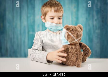 Kleiner Junge, der mit einem Teddybären am Tisch sitzt In medizinischen Gesichtsmasken Stockfoto