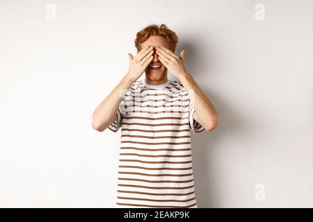 Lächelnder Mann bedeckt die Augen und wartet auf Überraschung, steht in T-Shirt vor weißem Hintergrund Stockfoto