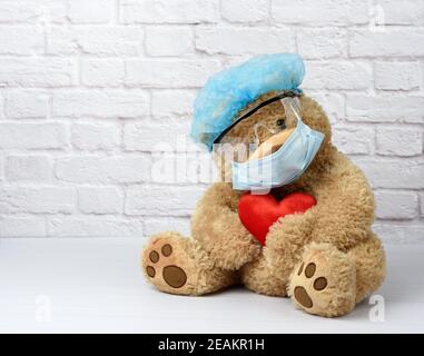Brauner Teddybär sitzt in einer Schutzbrille aus Kunststoff, einer medizinischen Einwegmaske und einer blauen Kappe Stockfoto