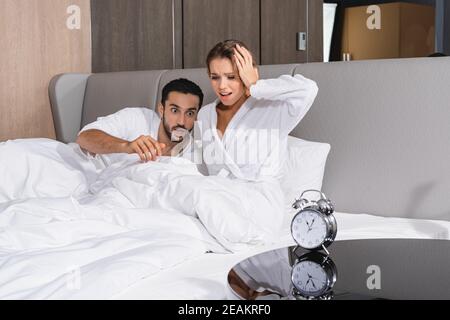Besorgt interrassisches Paar auf dem Bett in der Nähe des Weckers im Hotel Zimmer Stockfoto