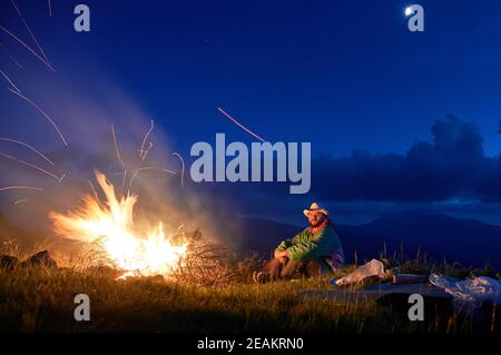 Männliche Camper sitzt in der Nähe hell orange Funken Flamme des Lagerfeuers in Bergräumen. Tourist entspannen am Lagerfeuer unter blauem bewölktem Himmel in kühler Sommernacht. Stockfoto