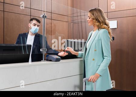 Lächelnde Frau mit Gepäck, die den Schlüssel vom Hotelmanager einnahm Medizinische Maske in der Lobby Stockfoto