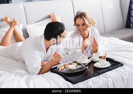 Fröhliche Frau im Bademantel Blick auf muslimische Freund mit Gabel Fast leckeres Frühstück auf dem Hotelbett Stockfoto