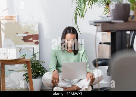 Glücklicher afroamerikanischer Architekt, der lächelt, während er auf dem Boden sitzt Laptop im Heimstudio auf verschwommenem Vordergrund Stockfoto