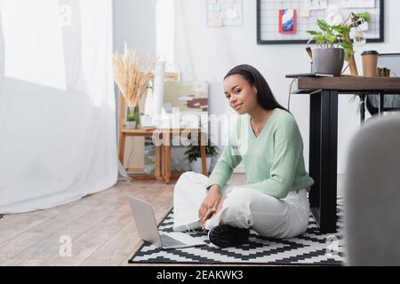 Junge afroamerikanische Architektin, die auf die Kamera schaut, während sie auf dem Boden mit Laptop sitzt, verschwommener Vordergrund Stockfoto