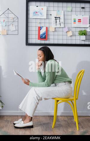 Junge afroamerikanische Innenarchitektin, die auf einem Stuhl sitzt und schaut In Skizzenbuch Stockfoto