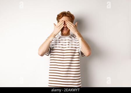 Lächelnder Mann bedeckt die Augen und wartet auf Überraschung, steht in T-Shirt vor weißem Hintergrund Stockfoto