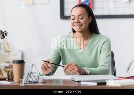 Glücklicher afroamerikanischer Architekt in Brillen, der Bleistift hält, während er sitzt Am Schreibtisch auf verschwommenem Vordergrund Stockfoto