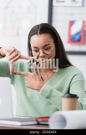 Lächelnder afroamerikanischer Architekt, der im Sitzen mit den Händen ein Quadrat zeigt Bei unscharfem Vordergrund Stockfoto