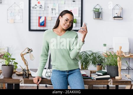 Glücklicher afroamerikanischer Architekt, der die Kamera anlächelt, während er an der Kamera mitteilst Smartphone Stockfoto