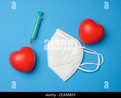 Rotes Herz und eine eingeklemmte Nadel einer Plastikspritze hinein, weiße Maske auf blauem Hintergrund Stockfoto