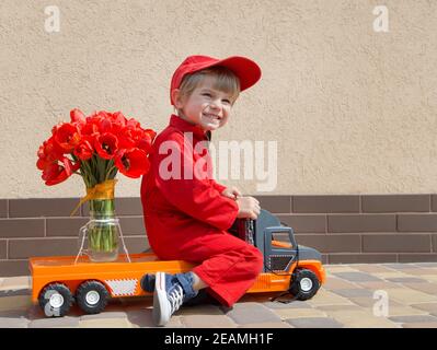 Lustige niedliche 4 Jahre alten Jungen in einem roten Mechaniker Overalls und eine Kappe sitzt auf einem Spielzeug-LKW mit großen Strauß Tulpen. Lieferkonzept, kleiner Kurier, Stockfoto