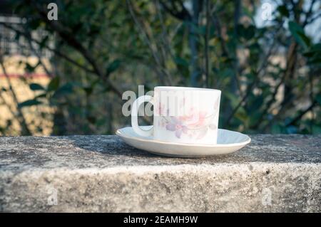 Kaffeetasse im Morgenlicht. Sommerlich frischer cooler Look. Weiße Kaffeetasse auf Untertasse für Heißgetränk auf Dachbalken eines Wohnhauses mit Bokeh Hintergrund. Stockfoto