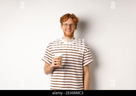 Hübscher junger Mann mit Bart und roten, unordentlichen Haaren, Brille mit gestreiftem T-Shirt, Kaffee zum Mitnehmen und lächelnd, weißer Hintergrund Stockfoto