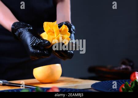 Reife Mango in den Händen des Küchenchefs. Der Koch schneidet Mango. Zubereitung von Mango- und Krake-Salat. Nicht erkennbare Person. Foto im Innenraum. Stockfoto