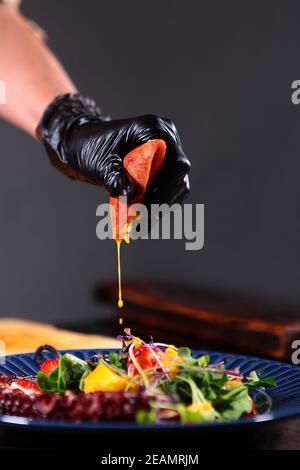 Der Koch gibt dem Oktopus und dem Mangosalat frischen Passionsfruchtsaft hinzu. Nicht fokussiert. Kochen der Zutaten für den Oktopus-Salat. Vertikales Foto. Stockfoto