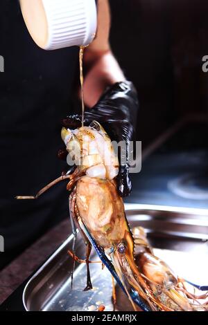 Der Koch gießt Sauce über riesige Süßwasser-Garnelen. Gegrillte Garnelen. Foto auf schwarzem Hintergrund. Vertikales Foto. Kochkonzept. Stockfoto