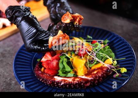 Der Koch fügt dem Krake-Salat Feigen hinzu. Hände außer Fokus. Octopus Salat mit Mango und Erdbeeren auf einem blauen Teller. Fotos ohne Personen. Kochkonzept. Stockfoto