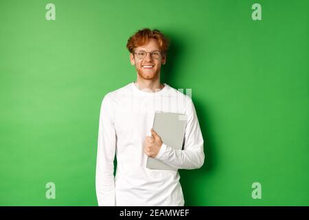 Hübscher junger Mann mit roten Haaren, Brille und Langarm-T-Shirt, Laptop auf grünem Hintergrund haltend Stockfoto