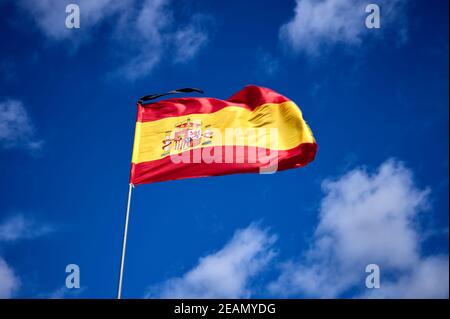 Detail eines Metallfahnenmastes mit einem roten und gelben Flagge von Spanien mit dem Wappen und einem Schwarze Kreppe an der Spitze gegen eine blaue und trübe Himmel Stockfoto