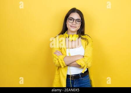 Junge Brünette Geschäftsfrau trägt gelben Blazer und Kopfhörer am Hals Auf gelbem Hintergrund mit einem fröhlichen Gesicht stehend und lächelnd Mit einem confide Stockfoto