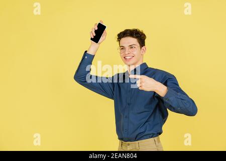 Zeigt auf leeren Bildschirm. Kaukasischen stattlichen Mann Porträt isoliert auf gelben Studio-Hintergrund mit Copyspace. Männliches Modell mit Telefon. Konzept der menschlichen Emotionen, Gesichtsausdruck, Verkauf, Werbung, Mode. Stockfoto