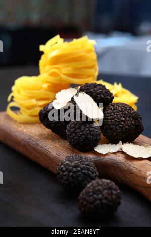 Schwarzer Trüffel auf einem Holzbrett mit Paste in Form von Nestern. Die Saison der Trüffel. Natürliches Antioxidans und Protein. Der teuerste Pilz. Vertikales Foto auf schwarzem Hintergrund. Foto im Innenraum. Stockfoto