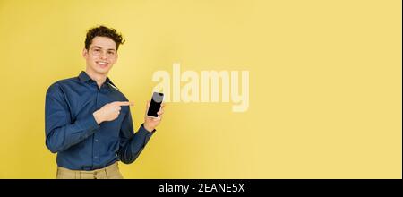 Zeigt auf dem Bildschirm. Kaukasischen stattlichen Mann Porträt auf gelben Studio-Hintergrund mit Copyspace. Männliches Modell mit Handy. Konzept der menschlichen Emotionen, Gesichtsausdruck, Verkauf, Werbung, Mode. Flyer. Stockfoto