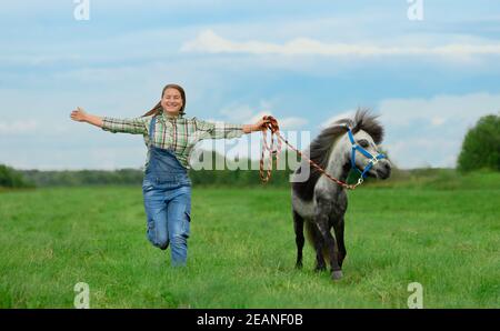 Lächelnde kaukasische Frau läuft mit weit ausgestreckten Armen. Erwachsene weibliche Trainerin im karierten Hemd und blauen Jeans und ihr Pony sind in ländlichen. Stockfoto