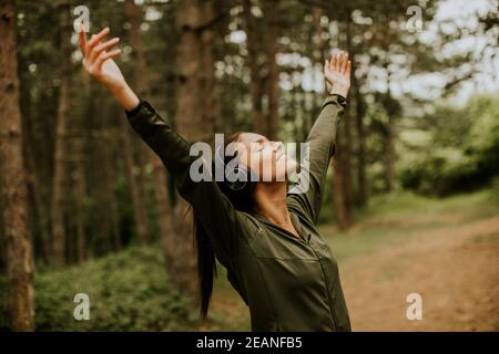 Hübsche junge Frau mit Kopfhörer, die ihre Arme im Wald vorpreading, weil sie draußen trainiert Stockfoto