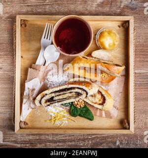 Mohn- und Apfelstrudel auf einem rustikalen Holztablett. Tee in einer Papiertasse mit einem weißen leeren Platz für Text und einem Löffel und einer Gabel aus umweltfreundlichen Materialien. Stockfoto
