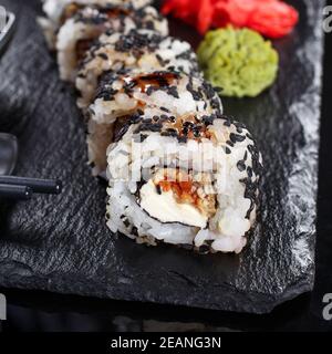 Traditionelle Japanische Küche. Sushi-Rolle mit Sesamsamen wird mit Essstäbchen und Sojasauce auf schwarzem Schiefer serviert, verziert mit Wasabi und Ingwer. Stockfoto