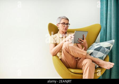 Stilvolle ältere Frau sitzt zu Hause auf einem Sessel und schaut sich Inhalte an Stockfoto