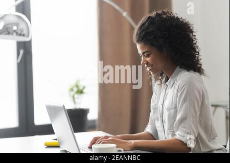 Seitenansicht Porträt einer positiv inspirierten afroamerikanischen Unternehmerin Geschäftsfrau, die am Schreibtisch sitzt und auf einem Laptop schreibt Stockfoto