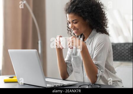 Lächelnde junge, fröhliche afroamerikanische Frau mit Afro-Frisur, die den Morgenkaffee genießt, während sie sich auf dem Laptop Videonachrichten und mit ihr spricht Stockfoto