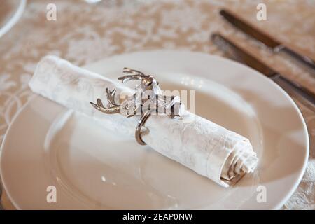 Die weiße Stoffserviette wird mit einem Ring in Form eines Hirschkopfes gesichert. Gabel und Messer aus dem Fokus.der Blick von oben. Makrofoto. Speicherplatz kopieren. Stockfoto