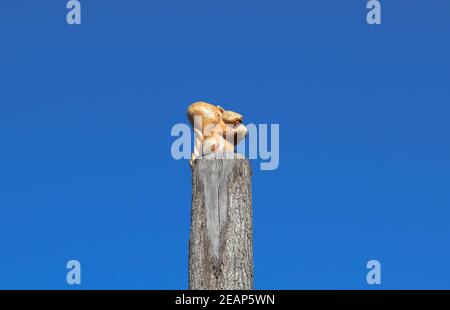 Die Figur des Eichhörnchens mit der Nuss vom Baum auf dem Baumstumpf auf dem Hintergrund des blauen Himmels. Figuren von Tieren aus Holz. Holzschnitzerei Stockfoto