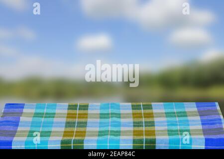 Leeres Tischprodukt. Leerer Holzdecktisch mit einer blauen karierten Tischdecke über abstrakt verschwommener natürlicher Kulisse. Platz für die Montage Ihrer Lebensmittel- und Produktdisplays. Stockfoto