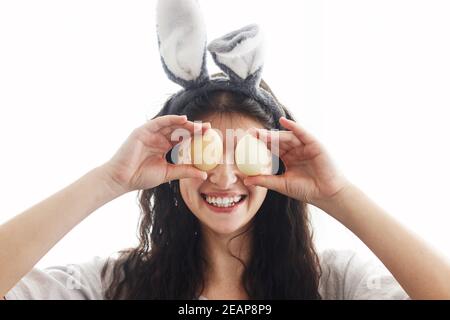 Frohe Ostern! Glückliche junge Frau in Hasenohren und Leinenkleid hält ostereier an den Augen und lächelt in weißem Licht. Natürlich gefärbte ostereier in der Hand Stockfoto