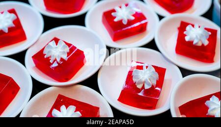 Rotes Gelee-Dessert mit Schlagsahne auf weißem Teller serviert. Quadratisches rotes Gelee auf dem Tisch des Restaurants für das Mittagsbuffet. Süßes Essen. Gelee-Agar. Dessert Buffet Catering Konzept, selektiver Fokus. Stockfoto