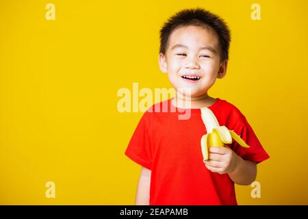 Kind niedlichen kleinen Jungen attraktives Lächeln trägt rote T-Shirt spielen Hält geschälte Banane zum Essen Stockfoto